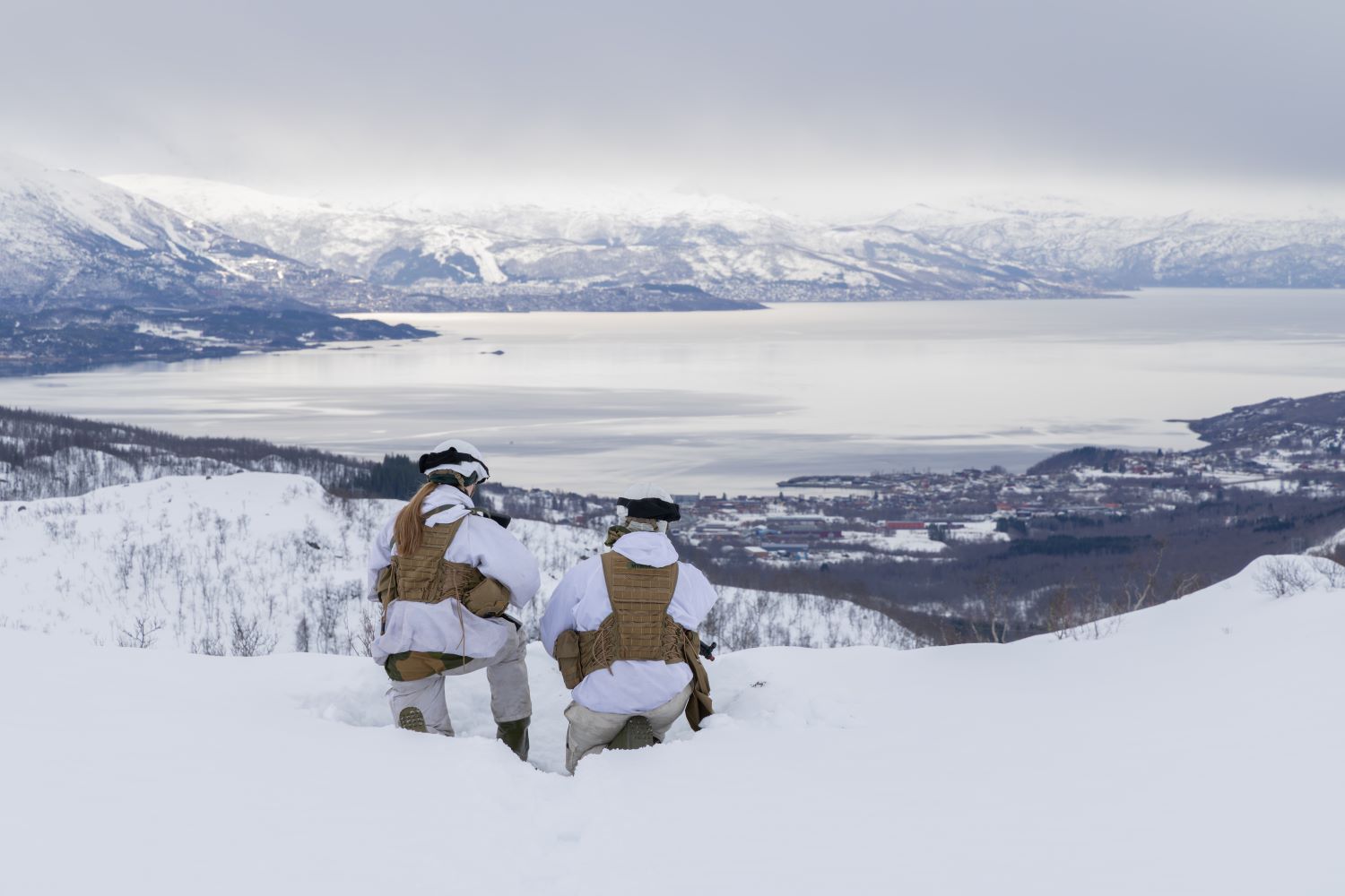 To soldater i vinteruniform med ryggen til i norsk vinterlandskap med fjell og fjord
